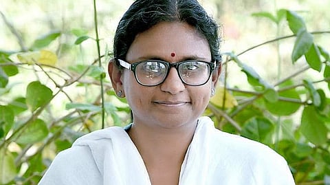 A medium shot of Meenakshi Natarajan, an Indian woman with a warm, gentle smile, looking directly at the camera. She has black hair parted down the middle and tied back, wears thick, black-rimmed glasses, and has a small red bindi on her forehead. She is dressed in a light blue-grey textured tunic (kurta) paired with a draped white cotton scarf (dupatta). The background features soft-focused, lush green foliage and thin branches outdoors in natural daylight.