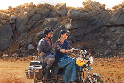 An image showing a smiling young man and woman riding a moped-style electric bike together in a rocky, arid landscape.
The woman is in the front, steering the moped while wearing a dark blue and brown traditional Indian outfit with her hair in a long braid. The man rides on the back, wearing grey overalls, boots, and a red-striped undershirt, smiling warmly at her. A yellow construction helmet hangs from the handlebars, and a large brown canvas bag is secured over the rear wheel. The background consists of massive, rugged grey boulders and piles of loose dirt under warm, golden sunlight.