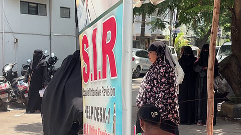 An outdoor street scene showing a help desk or registration stand, indicated by a prominent vertical banner with the large red letters "S.I.R." and smaller text below that reads "Special Intensive Revision" and "HELP DESK-1".
To the right of the banner, several women are lined up. One woman in the foreground faces left toward the banner, wearing a black hijab paired with a black and pink floral-patterned dress or tunic. Behind her in the queue, three other women stand completely clad in black niqabs and abayas. In the immediate foreground, the back of a woman's head with her hair tied up in a clip is visible.
To the far left, another woman in a black niqab walks near parked motor scooters against a white building wall. In the background, white cars are parked along a sunlit street with trees.