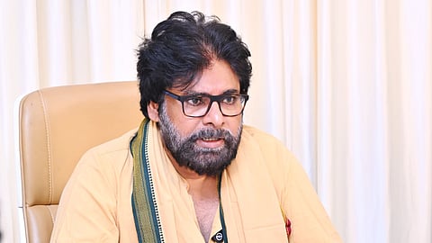 Pawan Kalyan with salt-and-pepper hair, a thick beard, and black-rimmed glasses sits at a dark wooden desk. He is wearing a light beige or peach-colored traditional long-sleeve tunic (kurta) with a green and gold bordered fabric sash (uttariyam) draped over his right shoulder.
He is looking slightly to his right with a focused expression, holding a pen in his right hand over some documents on the desk. In the foreground, a pen stand with various colorful pens, a small spray bottle, and an office conference speakerphone are visible. He is seated in a tan leather office chair against a backdrop of light cream-colored curtains.