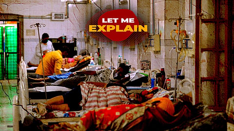 A gritty, high-contrast photograph of a crowded, dimly lit hospital ward with patients lying on close-packed beds, some with IV drips and oxygen masks. A healthcare worker in an orange shirt is seen attending to a patient in the background. The overall image has a textured, aged overlay with visible scratches and creases. Overlaid in the upper-center is a red textured badge with the bold text "LET ME EXPLAIN" in white and yellow.