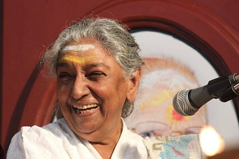 Veteran playback singer S Janaki smiles while seated beside a microphone during a public event, wearing a white sari and looking away from the camera.