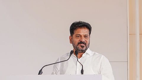 An Indian man with a beard and mustache, wearing a white button-up shirt, is standing behind a white podium and speaking into two microphones. The front of the podium features the official "amazon" logo with its characteristic yellow arrow. The background is a plain, light-colored wall with a vertical wooden panel visible on the right side.