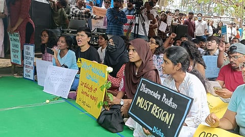 A passionate group of student protestors sits outdoors on a green tarp, chanting and holding signs. Prominent placards read "Dharmendra Pradhan MUST RESIGN!", "ERROR 404 ACCOUNTABILITY NOT FOUND", and "SCRAP NTA." Media personnel, an Indian flag, and determined faces fill the background of the demonstration.