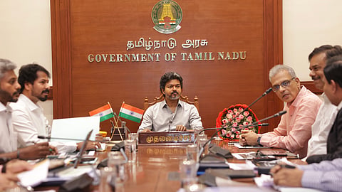 A medium wide shot of a government meeting room in Tamil Nadu, India. At the center of a long wooden conference table sits CM Vijay, looking forward with a serious expression. He is wearing a light grey button-up shirt. Behind him is a large wooden wall panel featuring the emblem of the Government of Tamil Nadu and the text "GOVERNMENT OF TAMIL NADU" in English and Tamil. In front of him on the table is a nameplate in Tamil that reads "முதலமைச்சர்" (Chief Minister). Several officials and individuals in formal attire are seated along both sides of the table, engaged in the meeting. Small Indian flags and a bouquet of pink flowers are also visible on the table.