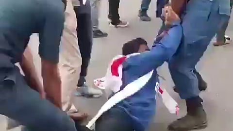 A photo showing security personnel in blue uniforms restraining an individual on the ground during an outdoors struggle. Two officers hold onto the person, who is wearing a blue long-sleeved shirt, dark pants, white sneakers, and a white and red sash. Other people and additional officers in light uniforms are visible standing in the background on a paved surface.