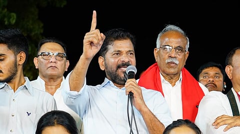 A candlelit rally featuring politician Revanth Reddy speaking into a microphone with his index finger raised, surrounded by other men on stage. In the foreground, two young women stand holding lit green candles—one wearing a black "OVER" hoodie and glasses, and the other wearing a black-and-white patterned top.