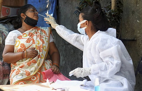 A healthcare worker wearing full protective gear, including a white PPE gown, surgical mask, and disposable gloves, collects a nasal swab sample from a woman seated outdoors. The woman being tested wears a pattern-printed sari and has her face mask pulled down below her chin, wincing slightly as the swab is gently inserted into her nostril. Test vials, paperwork, and hand sanitizer rest on the table in front of them.