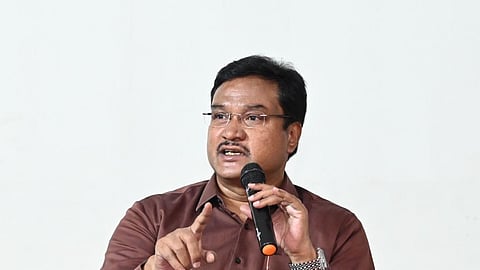 AV Ranganath with glasses and a mustache wearing a dark brown button-up shirt and grey trousers sitting on a stage, speaking into a microphone while pointing his index finger upward.