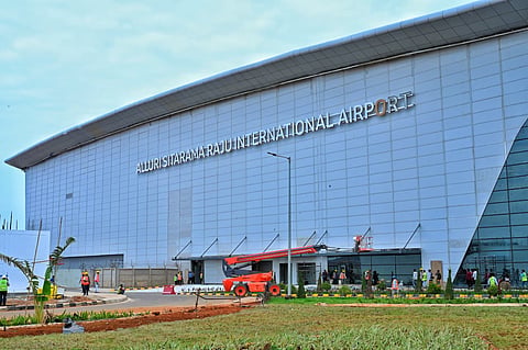 Exterior view of a modern, multi-story airport building under construction featuring light gray paneled cladding and large glass windows. Prominently displayed on the front facade in silver block lettering is the text "ALLURI SITARAMA RAJU INTERNATIONAL AIRPORT". An orange cherry picker crane/boom lift is parked near the ground-level entrance where several construction workers wearing safety vests and hard hats are walking and working. A paved road and landscaped grass area run along the foreground under an overcast sky.