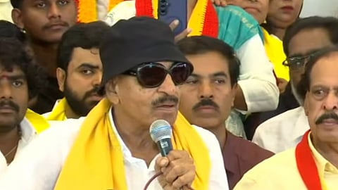 A close-up shot of Kannada activist and politician Vatal Nagaraj speaking into a microphone during a press conference. He is wearing his iconic black bucket hat, dark sunglasses, a white shirt, and a yellow and red scarf draped around his neck. Multiple news microphones with media logos are placed in front of him, and several supporters and attendees are visible sitting in the background.