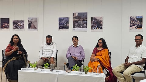 A panel discussion held at an indoor gallery event, featuring five panelists seated behind a white-draped table with small potted plants, water bottles, and glass tumblers. Name placards placed on the table in front of each speaker identify them from left to right:
Natasha Ramarathnam – A woman in a black and gold saree speaking into a microphone.
RV Karnan – A man in a white collared shirt and beige trousers looking down at his phone.
Jayesh Ranjan – A man in a light purple striped shirt and dark trousers seated centrally with hands clasped.
Anjali Lal Gupta – A woman in a bright yellow saree with a red border sitting attentive.
Prashanth Bachu – A man in a white short-sleeved button-up shirt and khaki trousers seated on the far right.
On the white wall behind the panel, several framed photographs depicting urban traffic congestion and cityscapes are hung in a row.