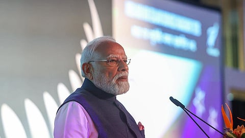 Prime Minister Narendra Modi stands at a podium, speaking into a pair of microphones. He is wearing spectacles, a light-colored long-sleeve shirt, and a dark navy blue Nehru jacket with a red pocket square. In the background, a large screen displays soft lighting and text, with a stylized silver leaf design on the left. A floral arrangement sits at the front corner of the podium.