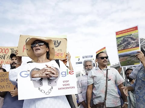 A group of activists holds demonstration signs during an outdoor protest against data center construction. In the center, a woman wearing sunglasses, a wide-brimmed straw hat, and a white shirt holds a sign featuring the Google logo with handcuffs chained across it and text that reads "STOP VIZAG DATACENTERS." Other protesters around her display additional signs with environmental messaging and images, including one titled "DATA CENTER RAMBILLI" showcasing deforested land.