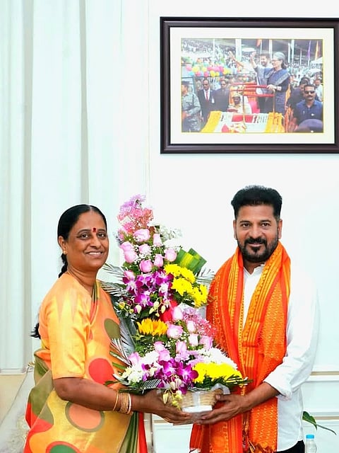 A woman in a bright orange saree with large yellow and green circles stands next to a man in a white kurta and an bright orange scarf around his neck. They are jointly holding a large, colorful floral arrangement filled with pink roses, purple orchids, and yellow sunflowers. A framed picture hangs on the light-colored wall behind them.