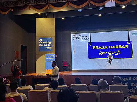 A speaker addresses an audience from a stage during a "Praja Darbar" event, with a large screen displaying the event title behind them, at a People's Convention on AI Data Centers in Vizag