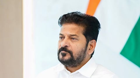 A portrait of Telangana Chief Minister A. Revanth Reddy sitting at a table with a microphone, wearing a crisp white shirt, with an Indian tricolor flag blurred in the background.