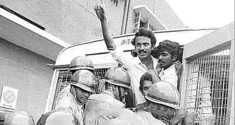 Black-and-white photograph of MK Stalin standing with other men near a police vehicle, raising one hand, with police personnel in helmets in the foreground.
