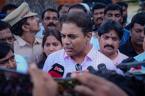 Indian politician K. T. Rama Rao wearing a light pink checkered shirt speaks to a crowd of reporters and microphones.