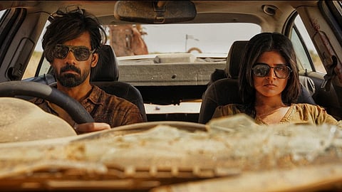 A still from 'Half'. A man and woman seated in a car. The man is in the driving seat. Both wearing sunglasses