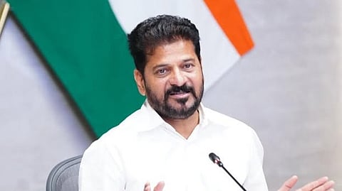 A portrait of Indian politician Revanth Reddy seated in front of an Indian flag background, wearing a white shirt and speaking with gestures.