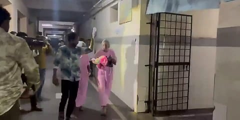 People, including medical staff in pink attire, carrying infants or bundles through a dimly lit hospital corridor beside a metal security gate.