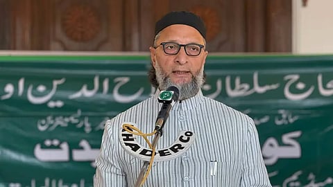 Asaduddin Owaisi, wearing glasses, a black skullcap (taqiyah), and a light gray striped kurta, speaking into a microphone with Urdu text on a green background behind him.
