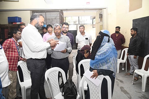 An official in a white shirt reviews documents held by another official while speaking to a woman wearing a blue headscarf and abaya during a field visit in an indoor setup.