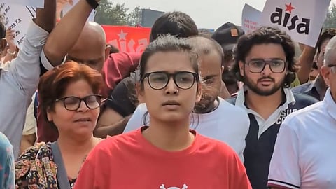 Neha Bora, wearing a red One Piece T-shirt and black-rimmed glasses, leads a group of protesters marching towards Jantar Mantar in Delhi. AISA flags and placards are visible among the crowd behind her.