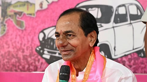 K. Chandrashekar Rao (KCR), leader of the Bharat Rashtra Samithi (BRS), speaking into a handheld microphone. He is dressed in a white kurta with a pink sash around his neck and is seated against a pink backdrop featuring a car graphic.