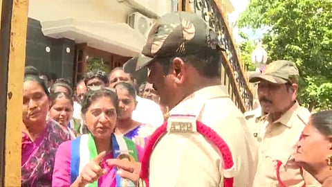 A woman in a pink saree wearing a party sash points her finger while speaking to a police officer standing outside a gate, surrounded by a crowd of people.