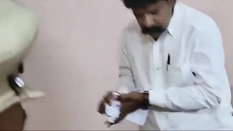 A man in a white collared shirt holding or tearing papers indoors while being observed by police personnel.
