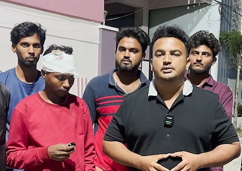 A man in a black polo shirt with a clip-on microphone speaks to the camera outdoors, accompanied by a group of men, including one with a white medical bandage wrapped around his head.