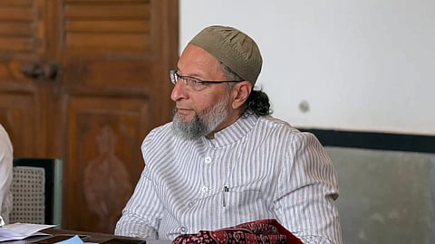 Asaduddin Owaisi seated indoors wearing eyeglasses, a brownish-green skullcap, and a light-colored pinstriped bandhgala jacket.