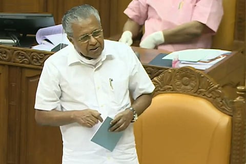 Senior CPI(M) leader and former Kerala Chief Minister Pinarayi Vijayan speaking inside the Kerala Legislative Assembly while standing near his seat. He is dressed in a white shirt and dhoti, holding papers in his hand during an assembly session.