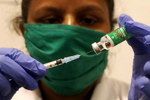 Medical professional wear green mask, holding a vial of vaccine and a syringe in her hands. She is wearing blue medical gloves.
