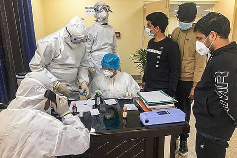 Four healthcare workers in PPE suits standing around a desk with three patients in masks beside them