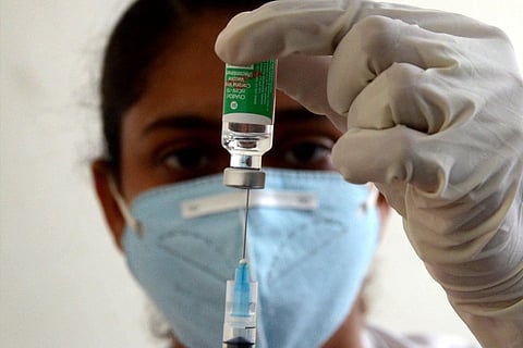 A health worker in a blue mask and gloves fills a syringe from a vaccine bottle