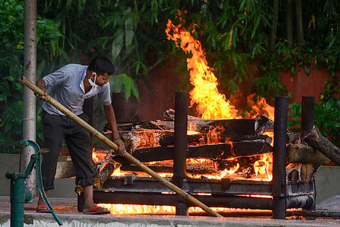 A man wearing a mask on his chin is seen moving the logs of a pyre where a body is burning