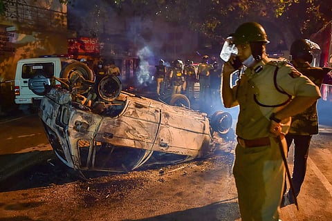A police officer stands on the left side of the picture talking over the phone. Behind him are the remains of an overturned vandalised jeep, which was set on fire.