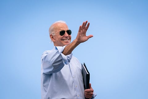 Joe Biden wears sunglasses and waves his right hand against a backdrop of blue