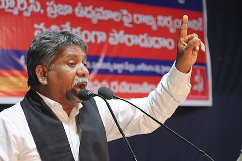 Manda Krishna Madiga is passionately speaking into a microphone at a public event. He is wearing a white shirt and a black scarf, and he gestures with his right hand, raising his index finger. Behind him is a red and blue banner with Telugu text, indicating a formal or political gathering. His facial expression and body language suggest that he is delivering an emphatic speech.
