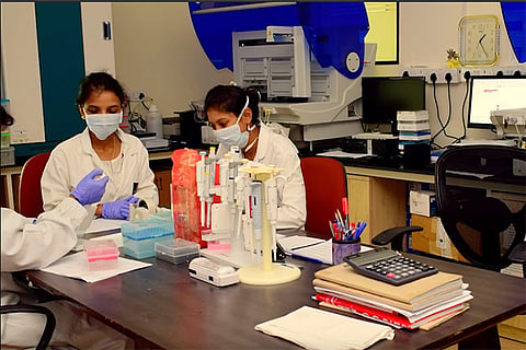 Three female lab technicians wearing masks and lab coats are working at a research lab table with pipettes, sample trays, and documents. The setting appears to be a virus research center, with lab equipment and computers in the background