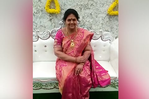 The statue of a woman sits on a white sofa wearing a pink and gold saree. Her hands are folded and she wears a smile on her face.