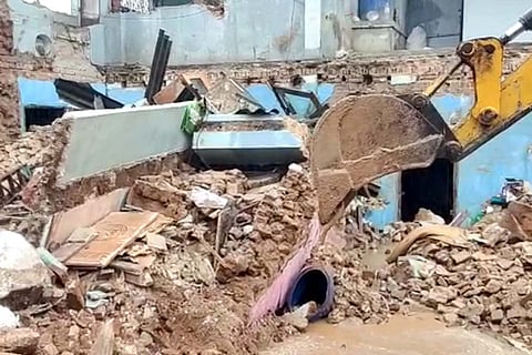 Rubble from a collapsed house with mud surrounding it and a JCB earth mover seen in the frame