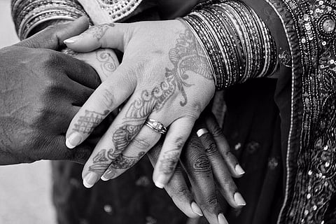 Black-and-white close-up of a couple’s hands layered together, adorned with wedding rings, bangles, and intricate mehndi designs, symbolising marriage and tradition. Image used for representation.