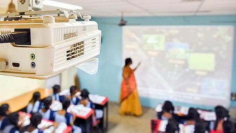 A photo of a classroom taken a ceiling height from behind a projector. Students can be seen in blue and white uniform. A teacher in an orange saree is explaining something on a large screen. The photo has been blurred to hide facial features.