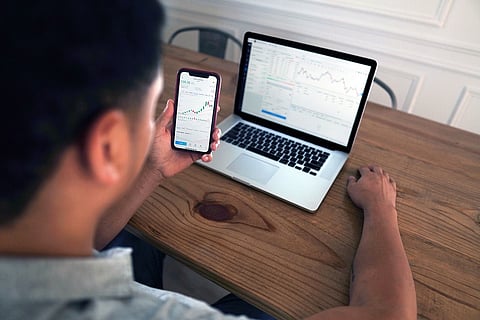 A man holds up a phone with stock market prices and a laptop is in front of him on a desk with trading info