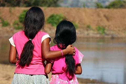 Two young girls wearing pink clothes, standing close with arms around each other, looking at a river — symbolising solidarity and the need for protection from child marriage.