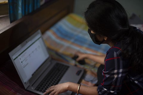 A woman in a plaid shirt wearing a mask and working on her laptop while sitting on a bed